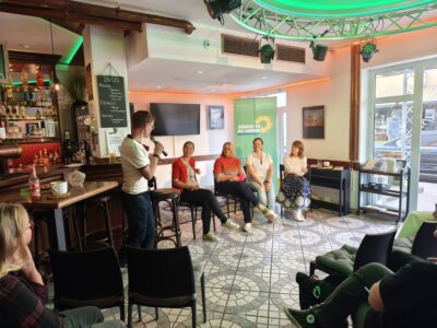 Blick in den Gastraum des Café Breddermann während der Podiumsdiskussion: Eine Frau steht mit Mikrofon vor der Bar und moderiert, vier Gästinnen sitzen auf Stühlen davor. Über der Runde hängt eine kreisförmige, grün beleuchtete Deckenkonstruktion mit Scheinwerfern, im Hintergrund sind Bar und ein Fenster zur Straße zu sehen